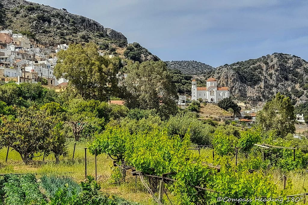 Town of white buildings along a hillside, with a lot of green gardens and vineyards around it. A handsome white church and the rugged mountains as a backdrop.