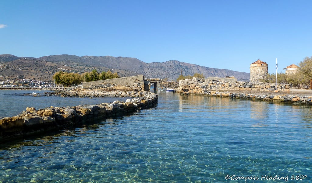 Clear turquoise water, stone walls and a bridge over the Elounda channel. Old windmills ashore.