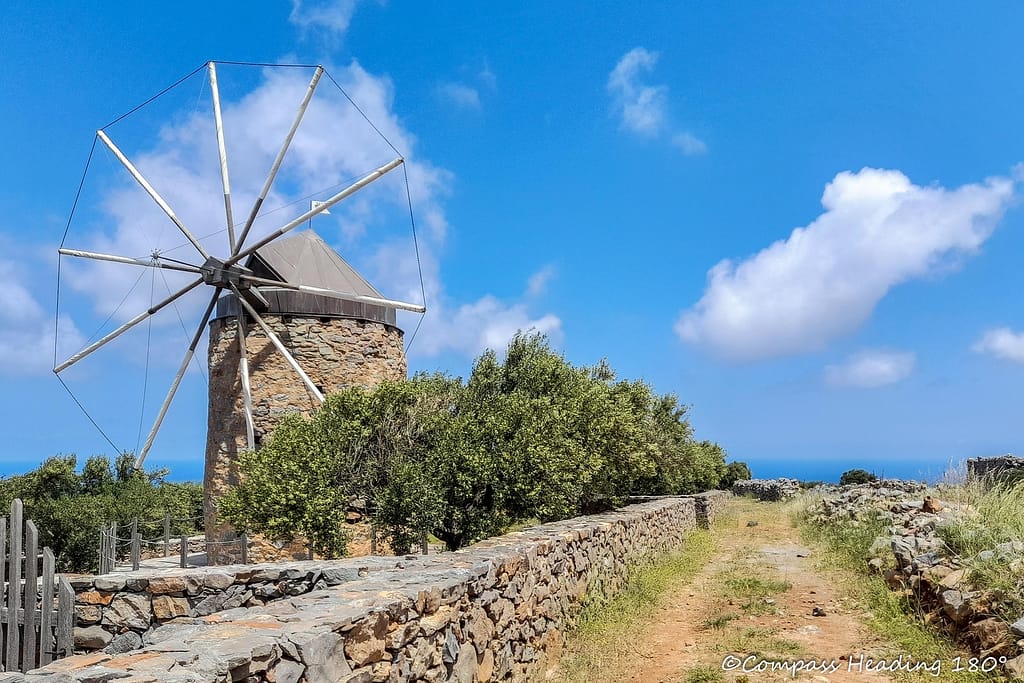 Small rounded windmill made of stone, a gravel road and the blue sea in the background.