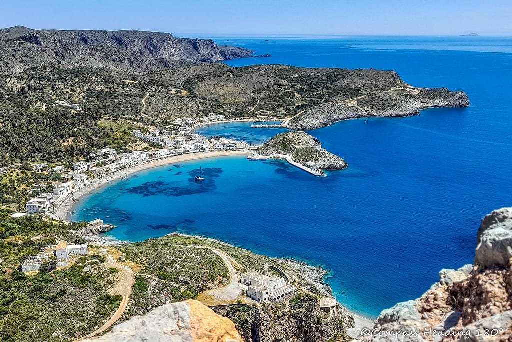 Kapsali bay of Kythira island, with only one boat in the middle, Aina. The anchorage is surrounded by a white sandy beach with a small village and hill all around. A smaller bay in the background is the local fishing harbour. The water is very clear, blue and turquoise.