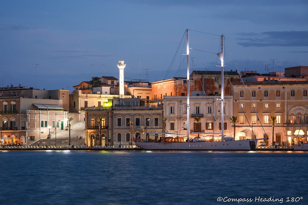 Night time scenery of the Brindisi old town, photographed from the opposite shore. Behind the seafront buildings the Roman column and large staircase, in front is a large sailing ship moored at the town quay.